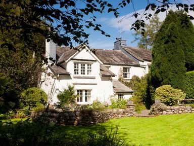 A white house with a slate roof surrounded by trees and a stone wall at High Cleabarrow in Windermere