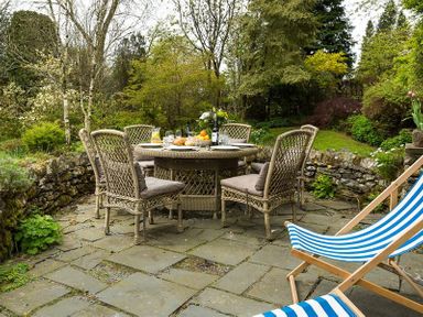 An outdoor stone patio with wicker chairs around a round table and striped deck chairs in a garden at High Cleabarrow in Windermere