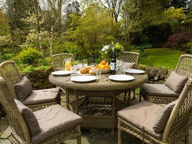 An outdoor dining area with a round wicker table and six cushioned wicker chairs set with plates glasses and food in a garden at High Cleabarrow in Windermere