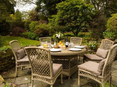 An outdoor patio area with a wicker table and six chairs set for a meal with plates drinks and flowers in a garden at High Cleabarrow in Windermere