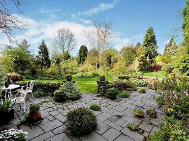 A stone patio with potted plants and white metal chairs and table overlooking a garden with grass and trees at High Cleabarrow in Windermere