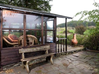 A wooden bench outside a glass enclosed sunroom with wicker chairs and a chiminea on a stone patio at High Cleabarrow in Windermere