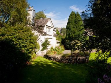 A garden area with shrubs and trees surrounding a white house at High Cleabarrow in Windermere