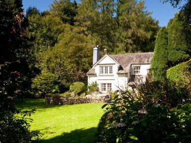 A white house surrounded by trees and bushes with a lawn in front at High Cleabarrow in Windermere
