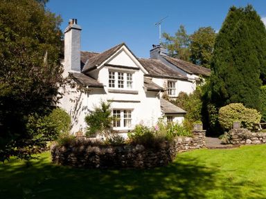 A white cottage with stone chimney and garden with stone walls and trees at High Cleabarrow in Windermere
