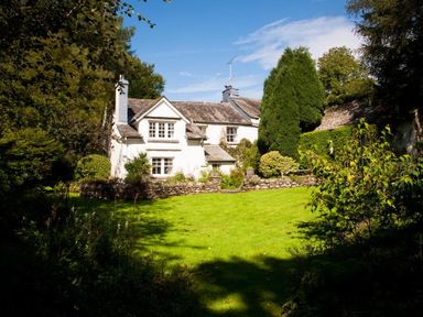 A stone house with chimney and green lawn surrounded by trees and bushes at High Cleabarrow in Windermere