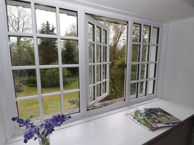 An open window with grid panes overlooking a garden with magazines and a vase of purple flowers on the windowsill at High Cleabarrow in Windermere