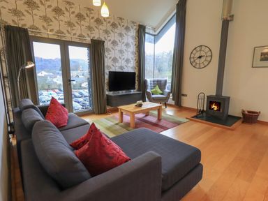 A living room with a grey sofa with red cushions a wooden coffee table a grey armchair a wall clock and a wood burning stove at Idle Mill 2 in Ambleside