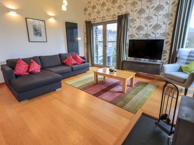 A living room with a grey sofa and red cushions a wooden coffee table on a striped rug a television on a stand floral wallpapered wall and large windows at Idle Mill 2 in Ambleside