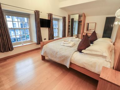A bedroom with a double bed, two large pillows, folded towels on the bed, two windows with curtains, a wall-mounted television, a mirror, and a wooden floor at Idle Mill 2 in Ambleside