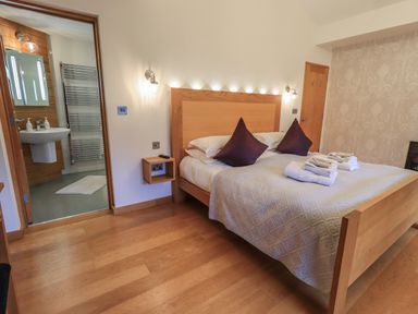 A bedroom with a wooden bed, purple pillows, folded towels, a wooden floor, wall lamps, and an en suite bathroom at Idle Mill 2 in Ambleside