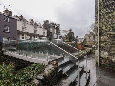A glass and metal pedestrian bridge over a small stream with stone buildings and houses in the background at Idle Mill 2 Ambleside