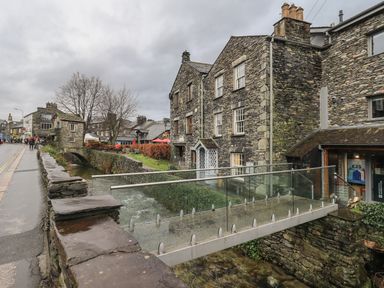 A stone building next to a small river with a glass pedestrian bridge and a street with people walking at Idle Mill 2 Ambleside