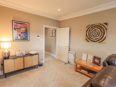 A living room with a wooden sideboard with a lamp and a framed picture a wooden coffee table with books and frames and a leather sofa at Waterhead Retreat in Ambleside