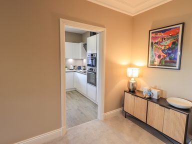 A hallway with a sideboard lamp and artwork leading to a kitchen with white cabinets at Waterhead Retreat in Ambleside