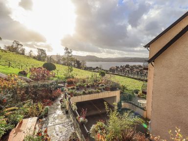 A garden with plants and a paved path next to a building overlooking a green field and a lake at Waterhead Retreat in Ambleside