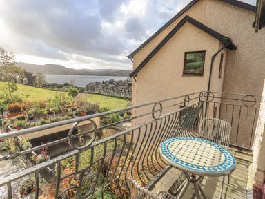 A balcony with a round table and two chairs overlooking houses a grassy area and a lake at Waterhead Retreat in Ambleside