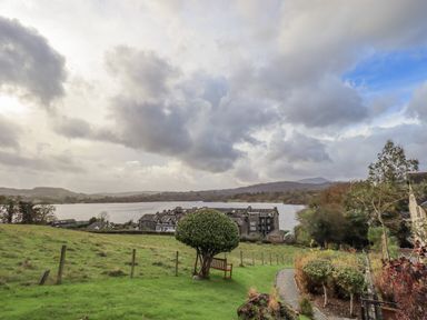 A grassy field with a trimmed tree and bench overlooking a row of houses and a lake with hills in the background at Waterhead Retreat in Ambleside