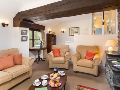 A living room with beige sofas and armchairs a wooden coffee table with tea sets and pastries a stone fireplace and wooden beam at Thirlmere Cottage in Grasmere