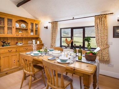 A dining room with a wooden table set with dishes and wine bottles near a window with floral curtains at Thirlmere Cottage in Grasmere
