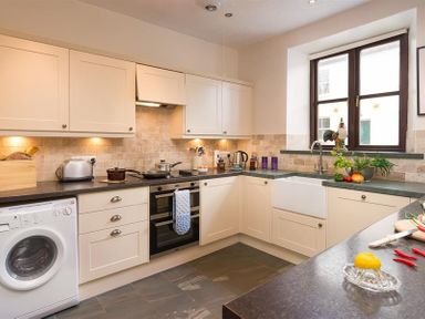 A kitchen with white cabinets black countertops a washing machine stove oven and a window at Thirlmere Cottage in Grasmere
