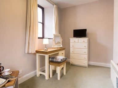 A room with a wooden dressing table with a lamp and mirror a matching stool with cushion and a white chest of drawers with a television on top at Thirlmere Cottage Grasmere