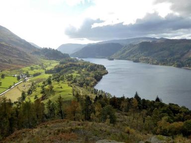 A lake surrounded by hills and forested areas with green fields and a road in the valley at Thirlmere Cottage in Grasmere