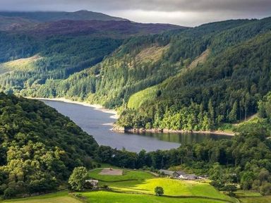 A river flowing between forested hills with green fields and houses in the foreground at Thirlmere Cottage Grasmere