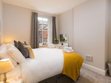 A bedroom with a bed covered with white and yellow bedding a window with gray curtains and a small dresser with a mirror at Thirty Blencathra Street in Keswick