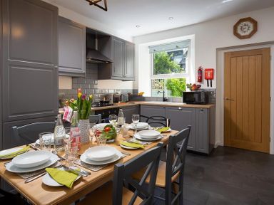 A kitchen with grey cabinets a wooden dining table set with plates glasses and cutlery at Fell Cottage Ambleside