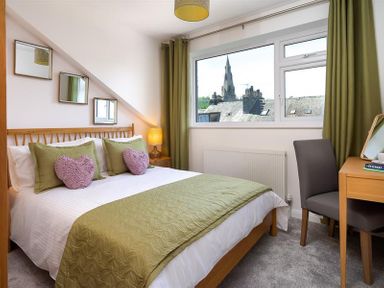 A bedroom with a double bed with green and pink pillows a bedside table with lamp a dressing table with a chair and a window showing rooftops and a church spire Fell Cottage Ambleside