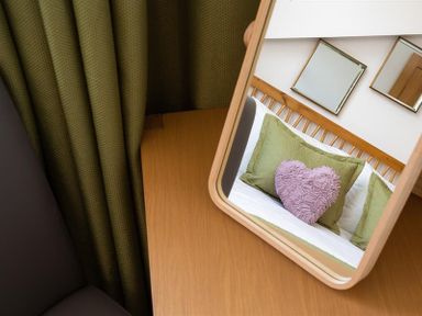 A mirror on a wooden table reflecting a bed with green pillows and a pink heart-shaped cushion at Fell Cottage in Ambleside