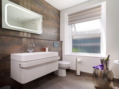 A bathroom with a wall mounted sink and faucet a mirror above and a toilet next to a window with a blind at Fell Cottage in Ambleside
