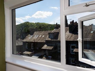 A view of rooftops and a green hill through a white-framed open window at Fell Cottage in Ambleside
