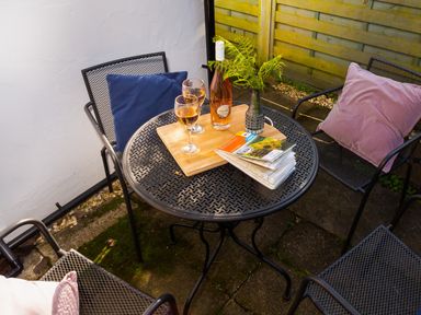 A round metal table with a bottle of wine two glasses a wooden tray a vase with ferns and maps on a patio with metal chairs and cushions at Fell Cottage in Ambleside