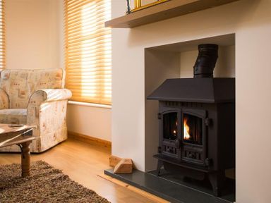 A room with a wood burning stove built into a wall next to a patterned armchair and wooden coffee table at Agathi in Keswick