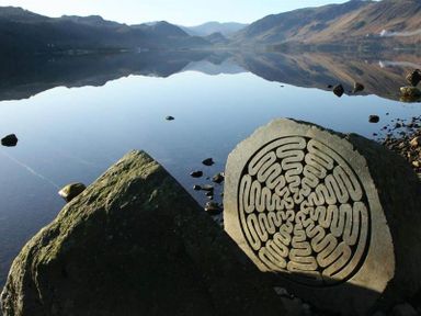A lakeside scene with mountains reflected in the calm water and a rock with a carved circular pattern by the shore at Agathi in Keswick