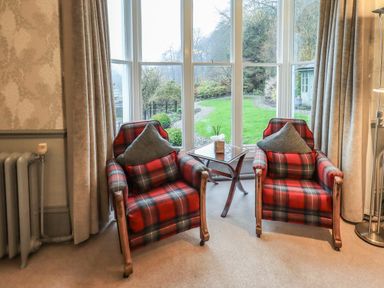 Two red plaid armchairs with cushions beside a small glass table in front of a large window showing a garden at Stepping Stones House in Ambleside