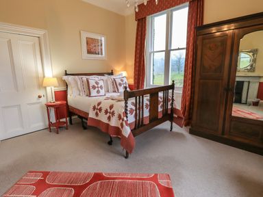 A bedroom with a wooden bed with patterned bedding a side table with a lamp a large window with red curtains and a wooden wardrobe with mirror at Stepping Stones House in Ambleside