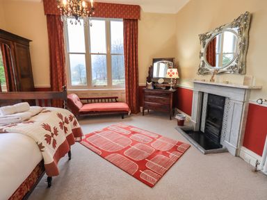 A bedroom with a bed covered by a patterned quilt a chaise lounge near a large window a wooden dresser with a mirror a fireplace with a large decorative mirror above and a red patterned rug on the floor at Stepping Stones House in Ambleside