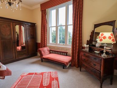 A bedroom with a wooden wardrobe a chaise lounge a wooden dresser with a mirror and a lamp and red curtains at Stepping Stones House in Ambleside
