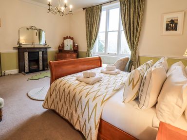 A bedroom with a wooden bed with pillows and towels a fireplace a dresser and large windows with curtains at Stepping Stones House in Ambleside