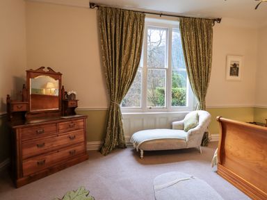A bedroom with a wooden dresser and mirror a lounge chair and green curtains at Stepping Stones House in Ambleside