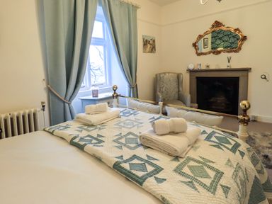 A bedroom with a bed featuring folded towels a patterned quilt curtains a chair a fireplace and a mirror at Stepping Stones House in Ambleside
