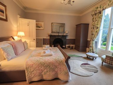 A bedroom with a bed linens and pillows a fireplace a wardrobe a window with floral curtains and circular rugs at Stepping Stones House Ambleside