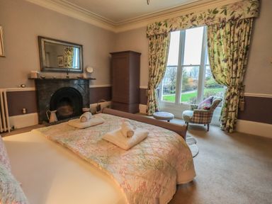 A bedroom with a bed a fireplace a wardrobe a window with floral curtains and an armchair at Stepping Stones House Ambleside