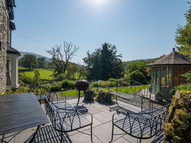 An outdoor patio area with metal chairs and tables overlooking green fields and trees at Stepping Stones House in Ambleside