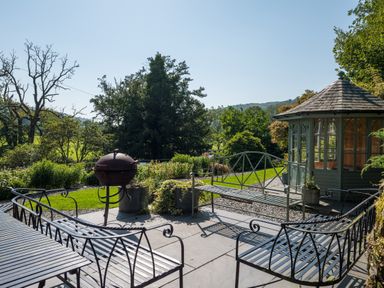 An outdoor patio with metal benches a metal grill and a gazebo surrounded by trees and plants at Stepping Stones House in Ambleside