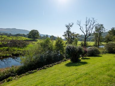 A grassy garden with bushes and trees near a river with hills in the background at Stepping Stones House in Ambleside