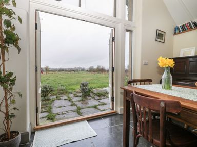 A dining area with wooden chairs and table with flowers in a vase and a view of a grassy garden through open double doors at Old Cary Cottage Henley near Langport